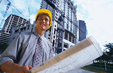 Photo of Asian male holding set of blueprints with yellow hard hat with commercial hi-rise building under construction in background.