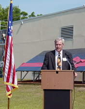 Photo: Philip Fairey speaking at podium in front of photovoltaic array with big red ribbon.